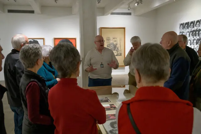Inside a gallery space with white walls, a museum educator stands behind a glass display case. He is wearing a white shirt, blue jeans, and gesturing with his hands in an explanatory manner. A group of older adults is watching intently, crowded around the display case, facing away from the camera and towards the educator. On the walls behind him, color pop art portrait paintings of famous people from the 1950s and 60s are barely visible.