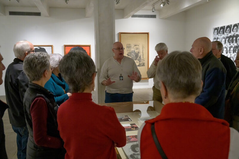 Inside a gallery space with white walls, a museum educator stands behind a glass display case. He is wearing a white shirt, blue jeans, and gesturing with his hands in an explanatory manner. A group of older adults is watching intently, crowded around the display case, facing away from the camera and towards the educator. On the walls behind him, color pop art portrait paintings of famous people from the 1950s and 60s are barely visible.
