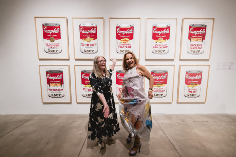 Two women celebrating in front of ten Wahol Soup Cans prints