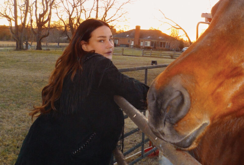 Woman with long red hair in black duster leaning on a fence with a horse muzzle in the foreground