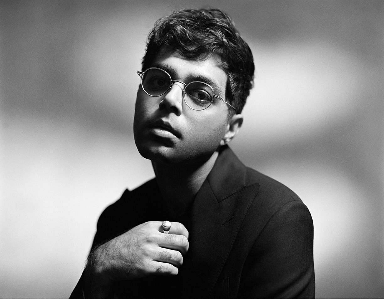 Portrait of Rafiq Bhatia wearing round glasses and a dark blazer, posing with his hand partially visible near their chin.
