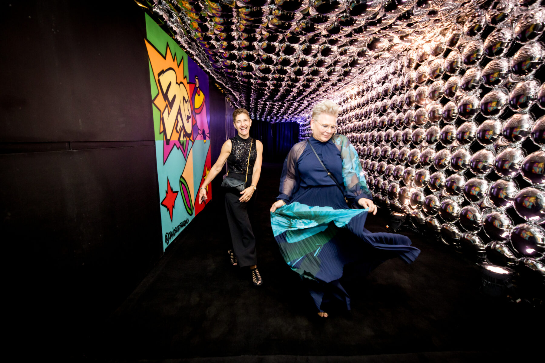 Two women, center image, walk down a black and silver hall