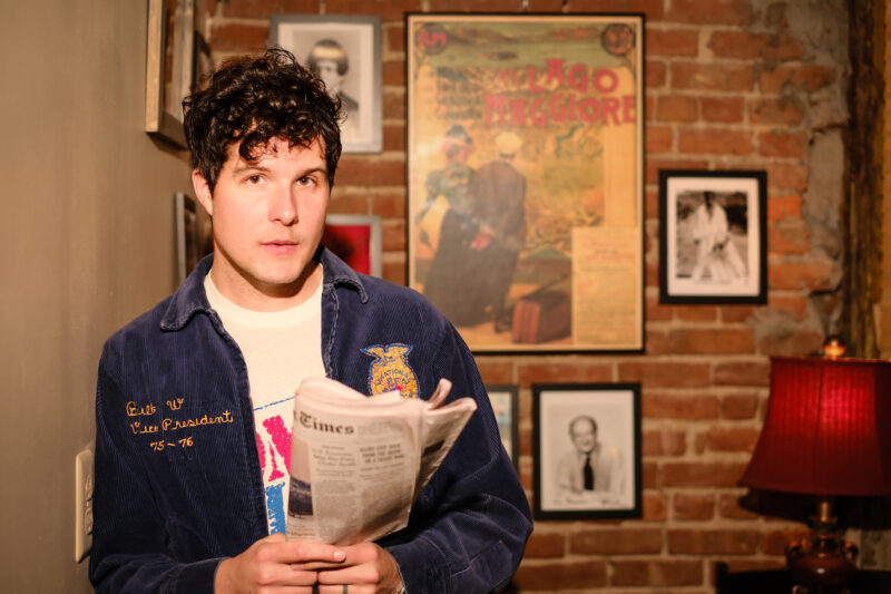 A young man with dark hair in a denim jacket holding a newspaper.