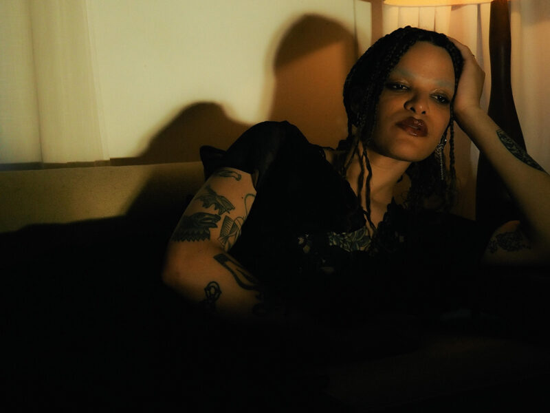 A person with braided hair and tattoos sits pensively in a dimly lit room, casting shadows on the wall.