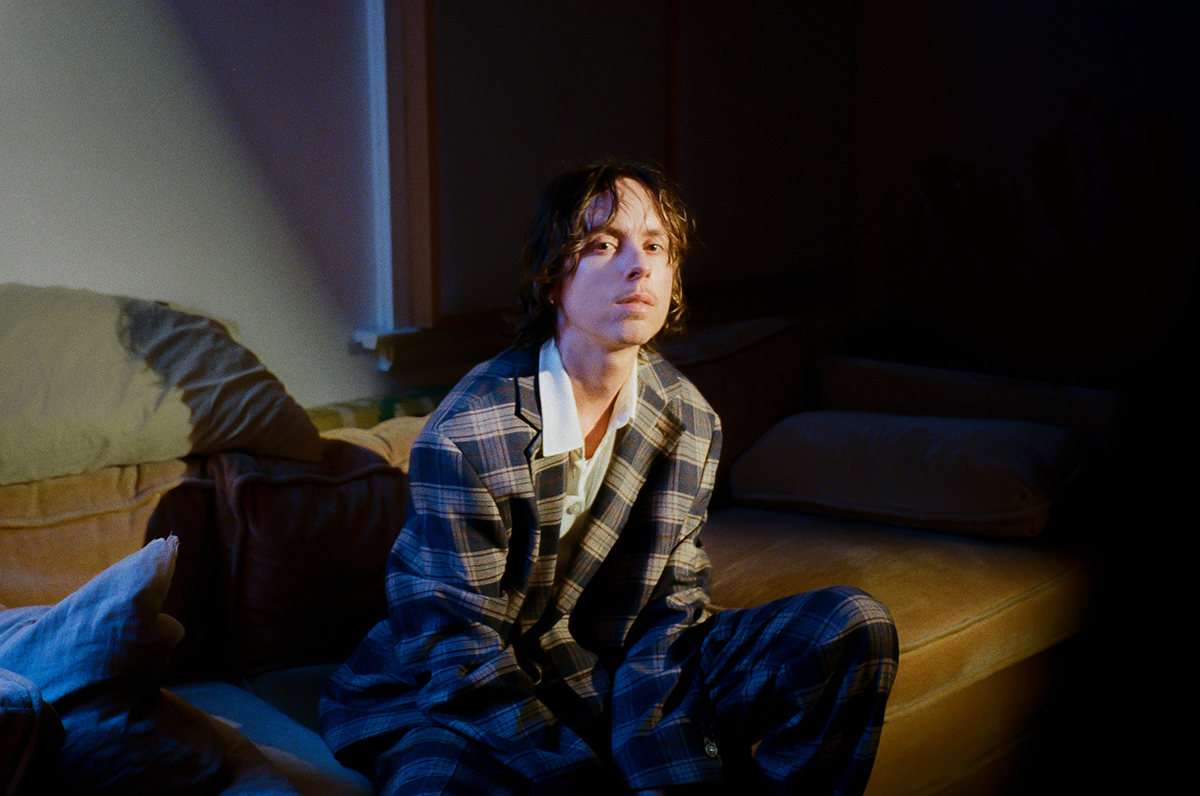 Person sitting on a mustard-colored sofa in a dimly lit room, wearing a plaid blazer and a thoughtful expression, bathed in soft natural light.