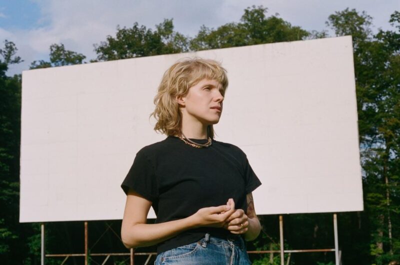 Person standing in front of a large, outdoor movie screen, surrounded by trees under a clear sky. The person is wearing a black T-shirt and sports a short, wavy hairstyle.