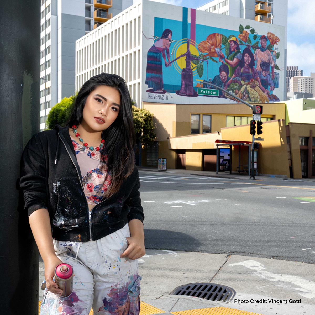 A person holding a spray paint can stands in front of a colorful mural with urban buildings in the background. There's a visible credit to photographer Vincent Gotti on the image.