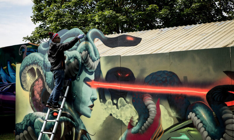 Artist on a ladder painting a large, vibrant mural featuring a fantastical creature with snake-like attributes.