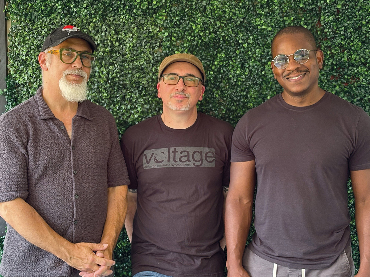 Three people standing side by side in front of a green hedge, the middle person wearing a 'Voltage' T-shirt.
