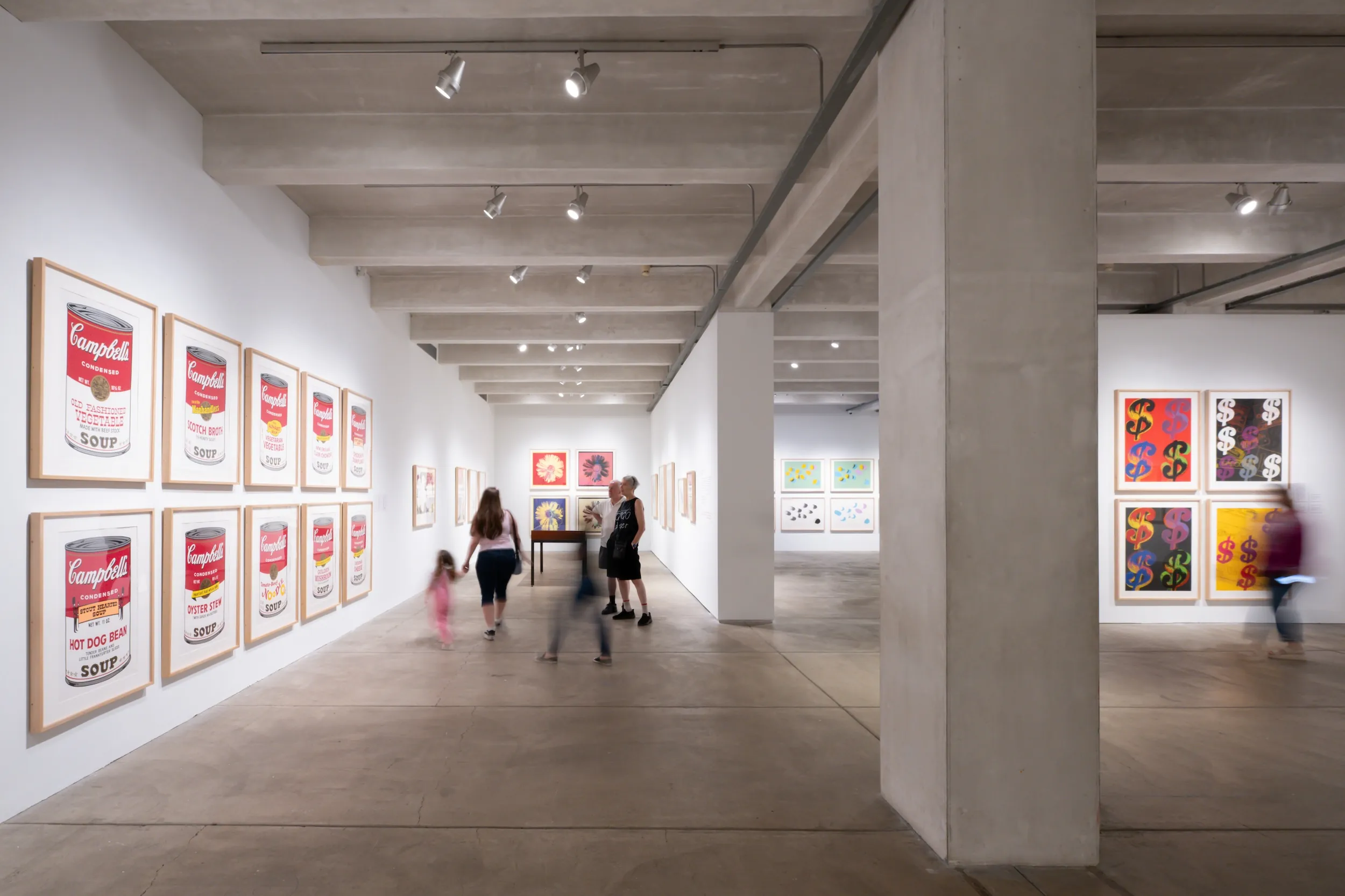 The image shows an art gallery with several framed artworks on the walls. On the left wall, there are multiple framed prints of Campbell's Soup cans in various colors and flavors. In the center of the gallery, a group of people, including adults and children, are walking and observing the artworks. The right wall features colorful prints with dollar signs and other abstract designs. The gallery has concrete floors and ceilings, illuminated by track lighting.