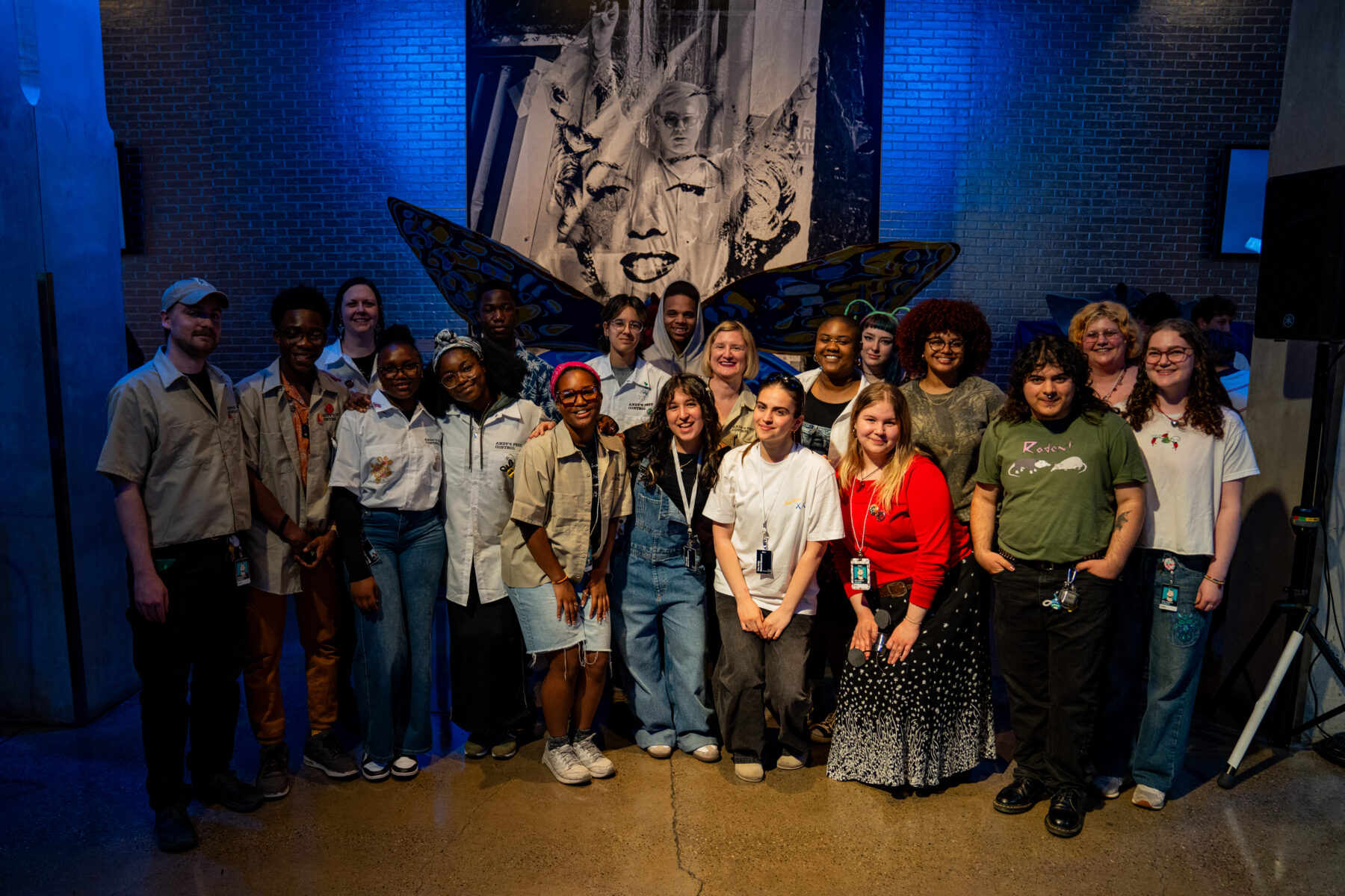 A group photo of 19 people stranding in front of a large painting of Andy Warhol and Marilyn Monroe