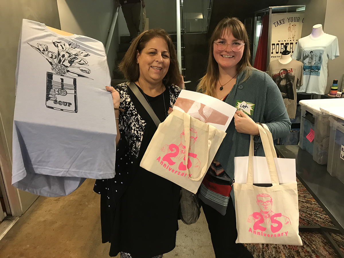 Two individuals smiling each holding up a tote bag commemorating the 25th Anniversary of The Andy Warhol Museum, one also displaying a T-shirt with a design.