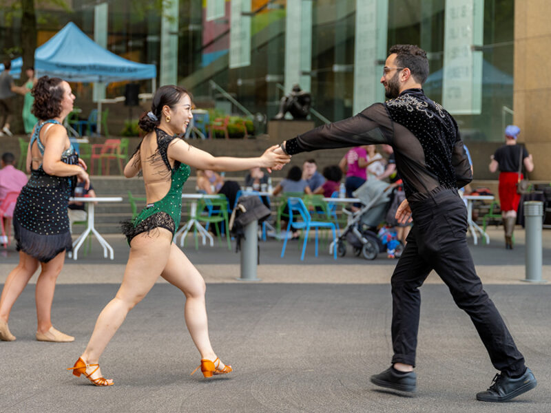 Two dancers in elaborate costumes performing a Latin dance at an outdoor event with spectators in the background.