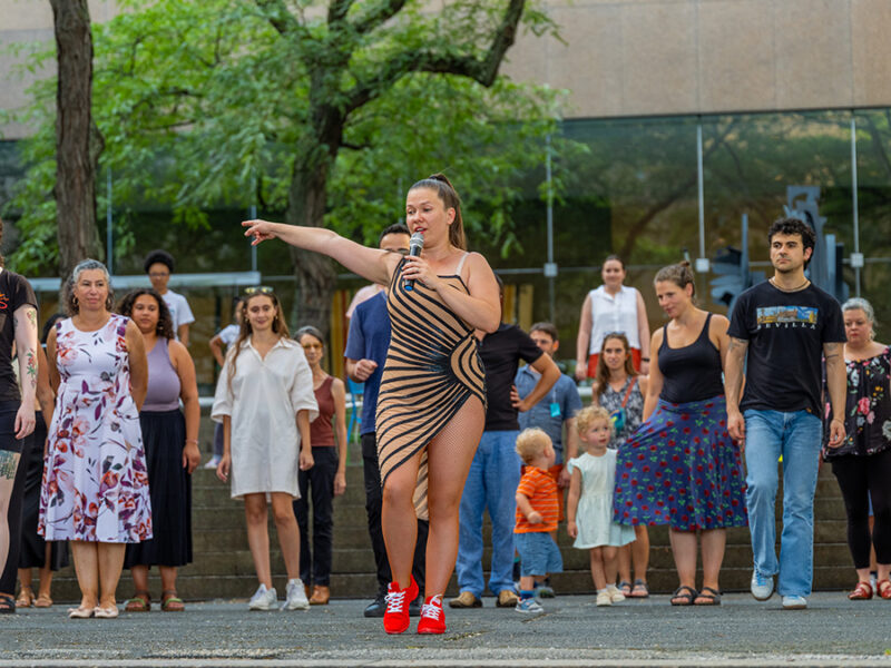 A person in a striped outfit and red shoes passionately singing and pointing, surrounded by an attentive crowd outdoors.