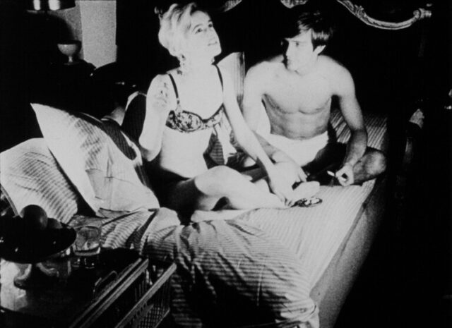 Black and white still from the film showing two characters sitting on a bed in conversation, one dressed in a bra and underwear and the other shirtless.