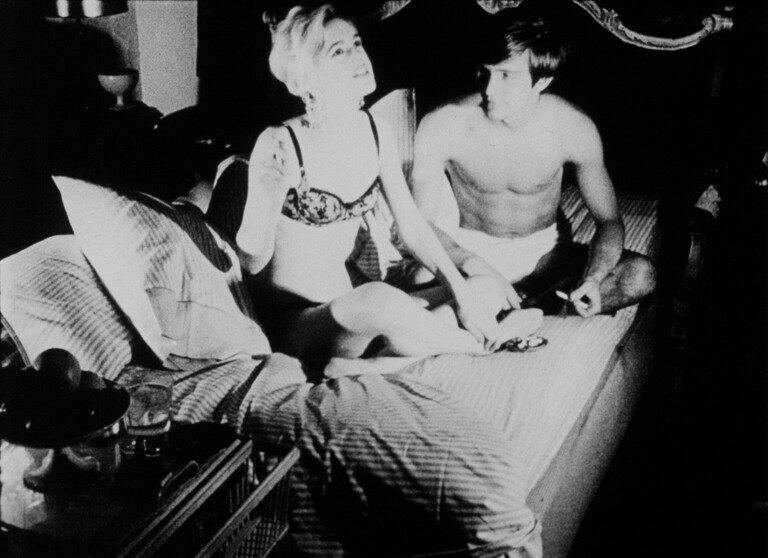 Black and white still from the film showing two characters sitting on a bed in conversation, one dressed in a bra and underwear and the other shirtless.