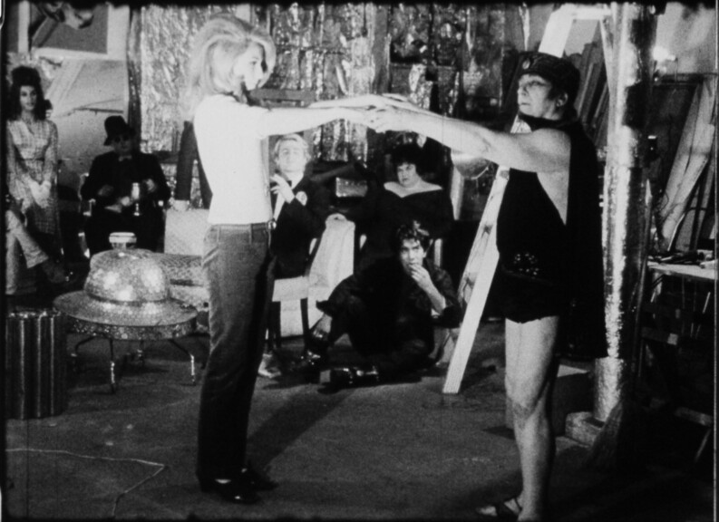 Film still of two people in a film scene inside the Silver Factory, with one person guiding the other's hand movement. Other observers are visible in the background amidst reflective decor and film equipment.