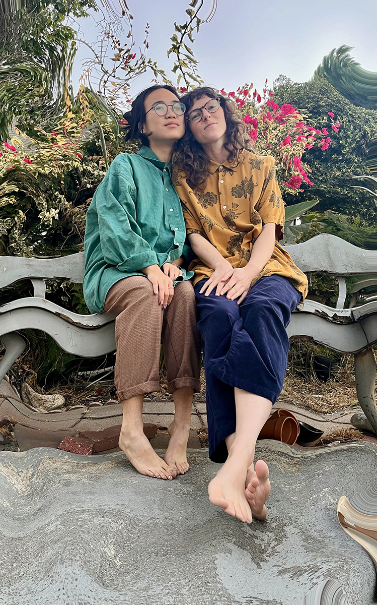 Two people sitting closely on a curved bench in a garden, with one resting their head on the other's shoulder. They are surrounded by lush plants and pink flowers. Both are wearing bright, casual shirts and glasses.