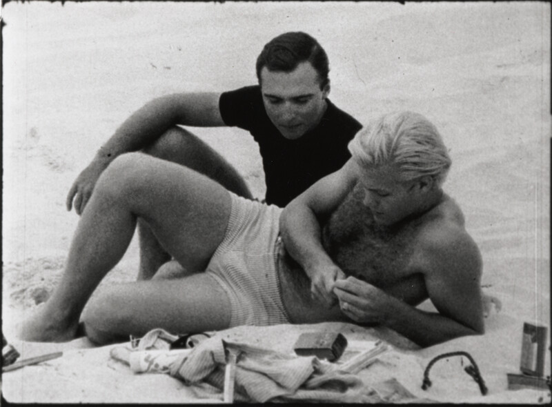 Black and white film still of a person sitting closely to another person on a sandy beach, both are engaged in a conversation.