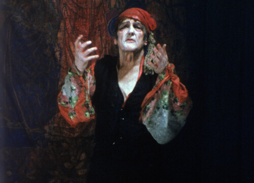Color film still of a person in theatrical makeup and vibrant costume performs emotively against a dark, draped background.
