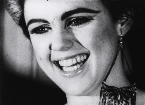 Black and white film still of a close-up of a smiling person with a stylish earring and an elegant necklace, expressing joy.