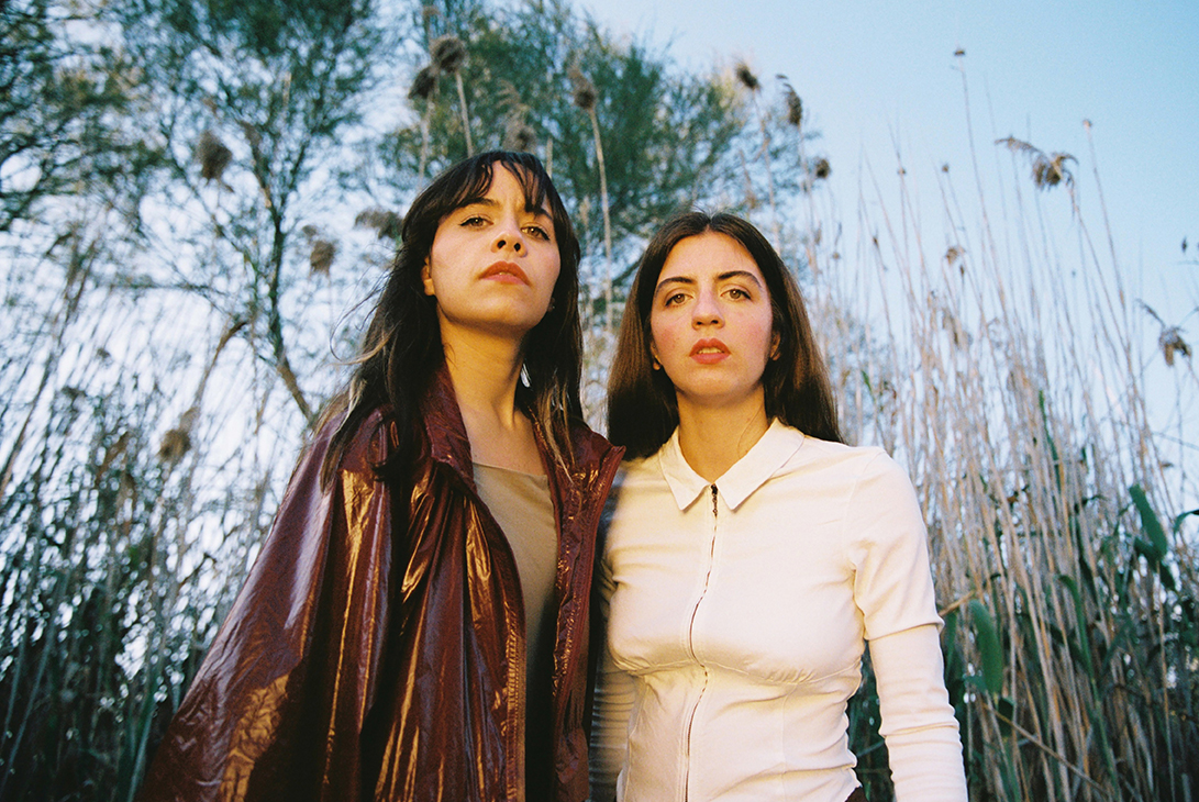 Two individuals standing in front of tall grasses with a clear sky in the background.