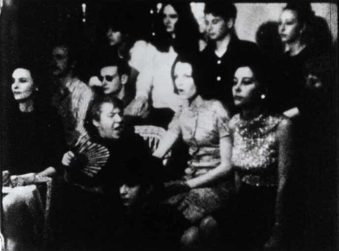 Black and white film still featuring a group of individuals sitting closely together, looking intently towards a focal point off-camera.