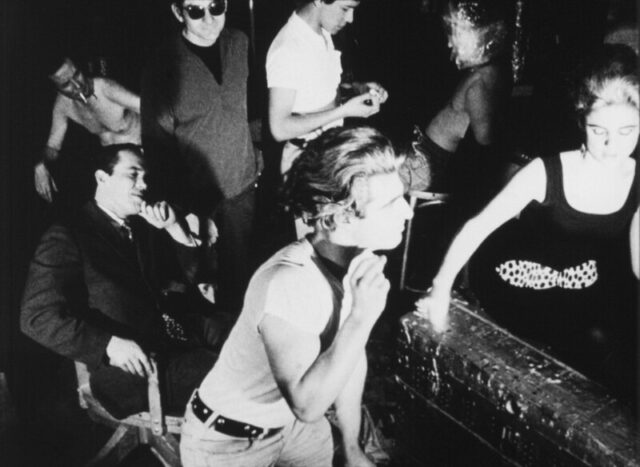 Black and white film still depicting a bustling scene as several individuals engage in various activities in a creative, energetic environment.