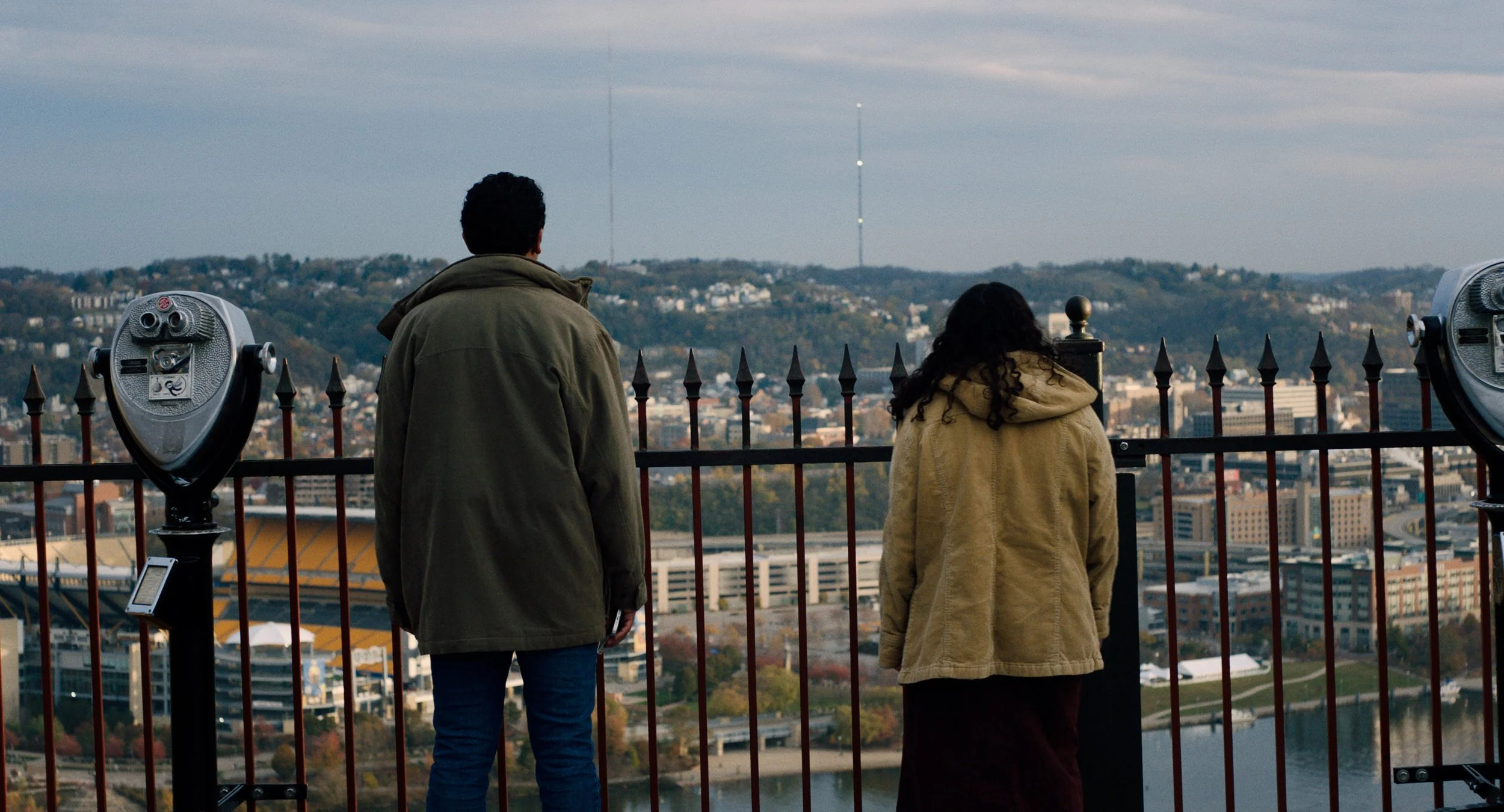 In a film still, two teenagers stand with their shoulders slumped and their backs to the camera. They are wearing fall coats and looking out over the city of Pittsburgh below them. The sky is overcast.
