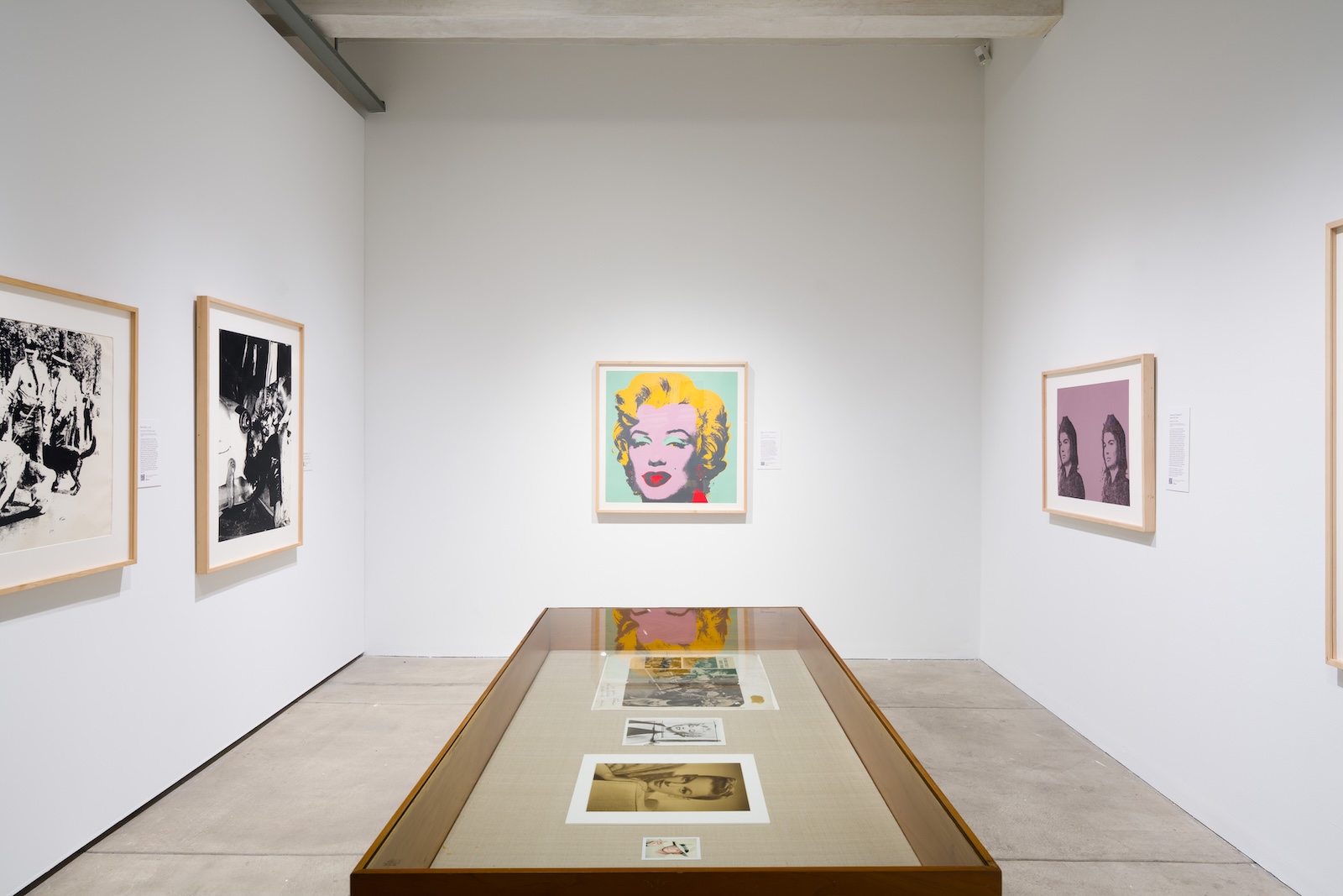 Art gallery room with white walls and a concrete floor. Framed artworks hang on all three visible walls. The left wall displays two black-and-white pieces. The back wall features a colorful artwork with a central rectangular area that is obscured. The right wall shows a framed piece depicting two figures. A glass display case in the center of the room contains various prints or photographs.