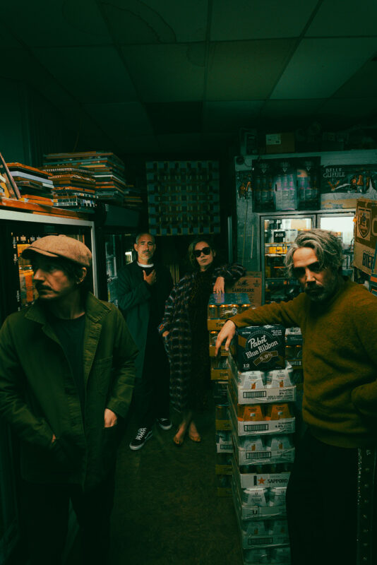 Four individuals posing stylishly in a dimly lit beer store, surrounded by shelves and stacks of beer cases.