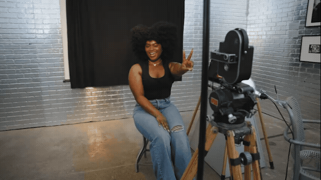 Person sitting on a stool in front of a camera setup, wearing a black tank top and ripped blue jeans, making a peace sign. Background features silver brick walls and framed pictures.
