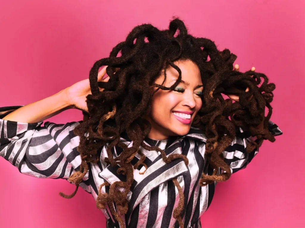 Person with voluminous curly hair smiling broadly, wearing a black and white striped jacket, against a pink background.