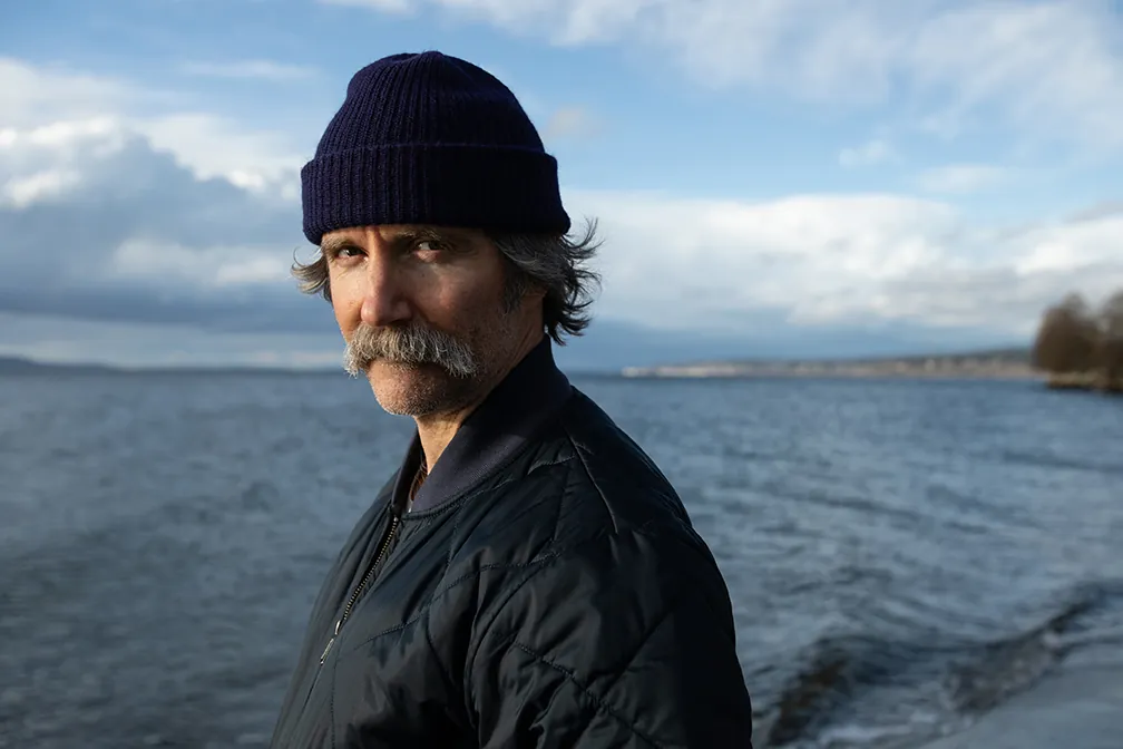 Person in a purple beanie and blue jacket standing by a lake with clouds in the sky.