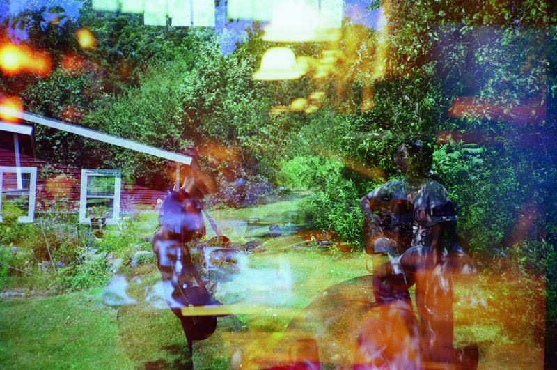 This photograph features a visually striking, abstract double exposure. It combines an indoor scene with a hanging lamp and outdoor view of a lush garden with a pathway and a small building. The colors are vibrant, dominated by green, blue, and splashes of orange.
