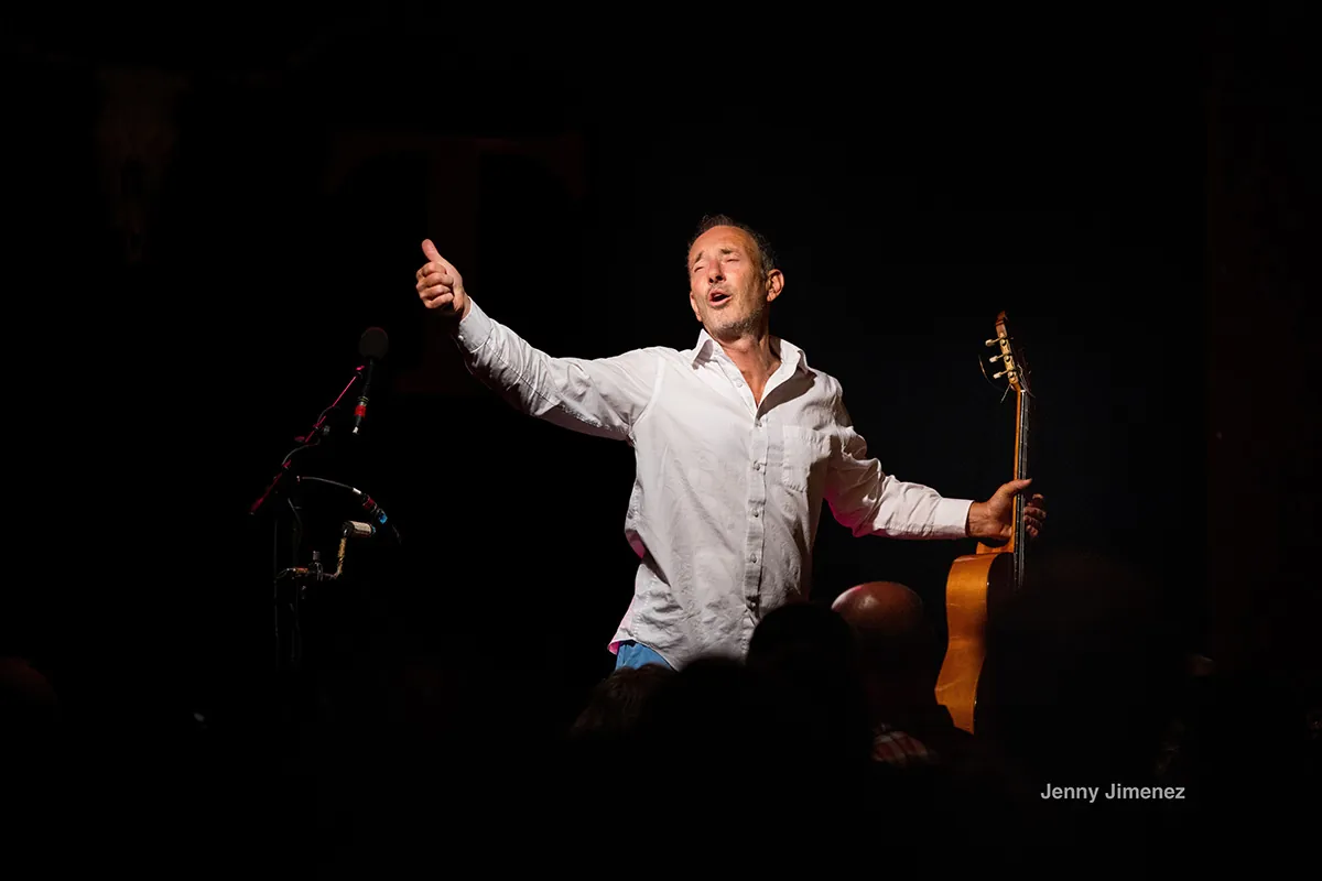 Musician Jonathan Richman performing on stage giving the thumbs up in one hand and holding a guitar in the other hand. A spotlight is illuminating him.