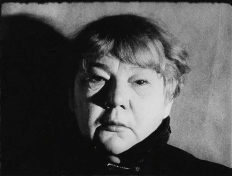 A film still of a woman's face. Strong light comes from her right, throwing the left side of her face in partial shadow. She has close cropped hair and stares at the camera with a neutral expression