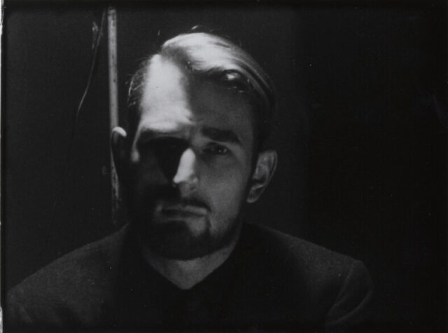 A film still of a man with a beard staring at the camera. Light comes down in a line over the right half of his head, pushing the left into relative shadow. His hair is combed over to the his left.