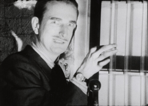 A black and white film still of a man in a suit to the left of center looking at the camera. He has a weird look in his eye, his mouth slighly open and the right corner raised. His had is level with his head, opening window shades.