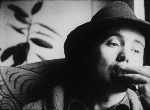 A black and white film still of a man, visible from the chest up in a fedora. He is putting food into his mouth, fingers just outside his lips, mouth wide open.