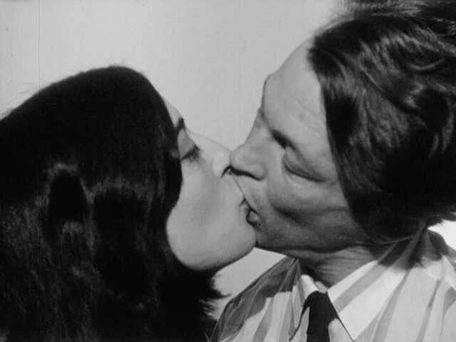 A black and white film still of a man and woman kissing. The camera views them from the side and both their heads are tilted in the kiss. The man's lips fully envelop the woman's.