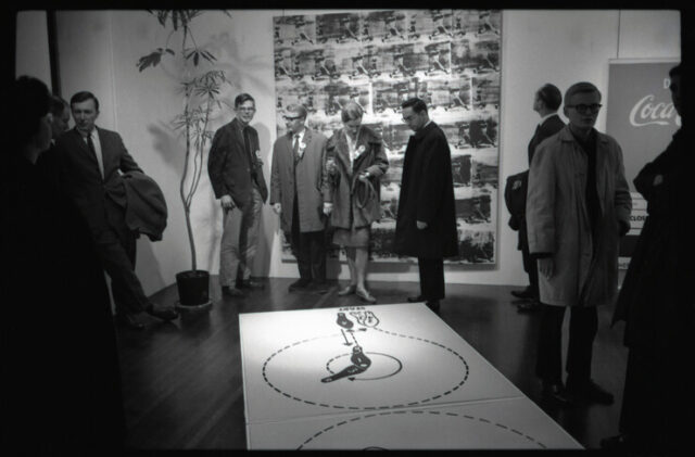 A black and white photo of well-dressed people standing in an art gallery. 10 people stand around a large painting on the floor with dance instructions.