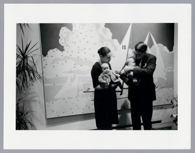 A black and white photo of a well dressed couple holding two babies in font of a large Do It Yourself painting of a sailboat, seemingly partially painted. Large potted ferns encroach from the corners of the shot.
