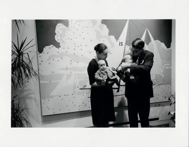 A black and white photo of a well dressed couple holding two babies in font of a large Do It Yourself painting of a sailboat, seemingly partially painted. Large potted ferns encroach from the corners of the shot.