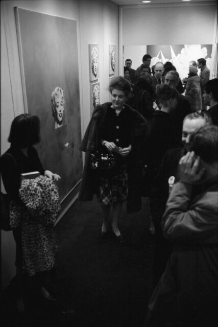 A black and white photo of a well dressed crowd of people at an art gallery opening. Center frame is a woman with a fur coat over her shoulders. On the wall are 5 Andy Warhol's Marilyn's.