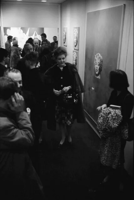A black and white photo of a well dressed crowd of people at an art gallery opening. Center frame is a woman with a fur coat over her shoulders. On the wall are 5 Andy Warhol's Marilyn's.
