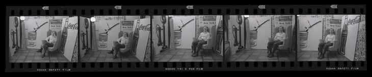 5 film negatives in black and white. Andy Warhol sits in a chair while surrounded by a paining of a Campbell's soup can and a Coca cola bottle. He shifts his stance slightly