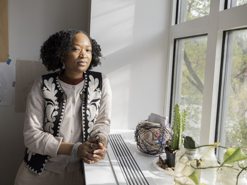 A woman leans against a window ledge looking at the camera. Light from the window shines across their face. Small plants sit on the window ledge.