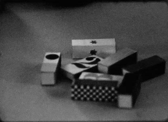 A black and white film still of 8 wood blocks scattered on the ground. Each block has it's own pattern.