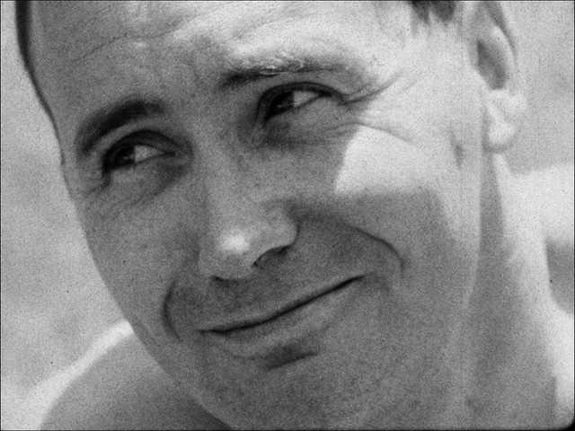 A black and white film still. A close of of a man faintly smiling, looking at something to his left. The small amount of bare shoulder we can see implies his shirtless.